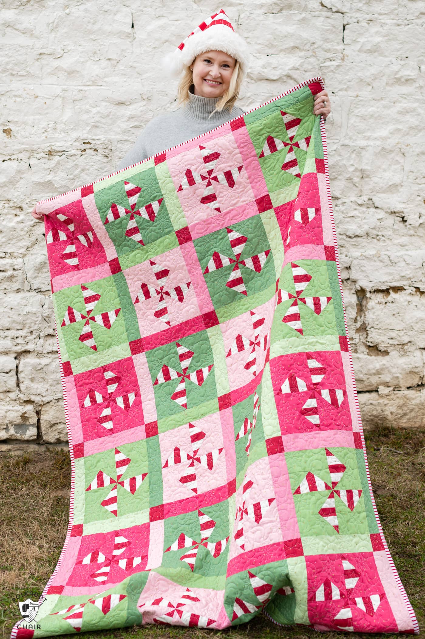 woman holding red, white, pink and green christmas quilt outdoors in front of a white brick wall