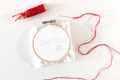 white fabric in embroidery hoop with red thread on a needle on white table